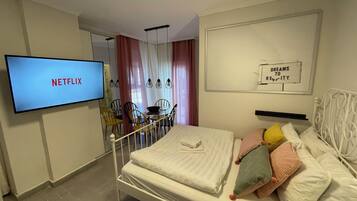A hotel room with a bed, a TV displaying 'NETFLIX', a 'DREAMS TO REITY' sign, and a dining area with chairs and a table.