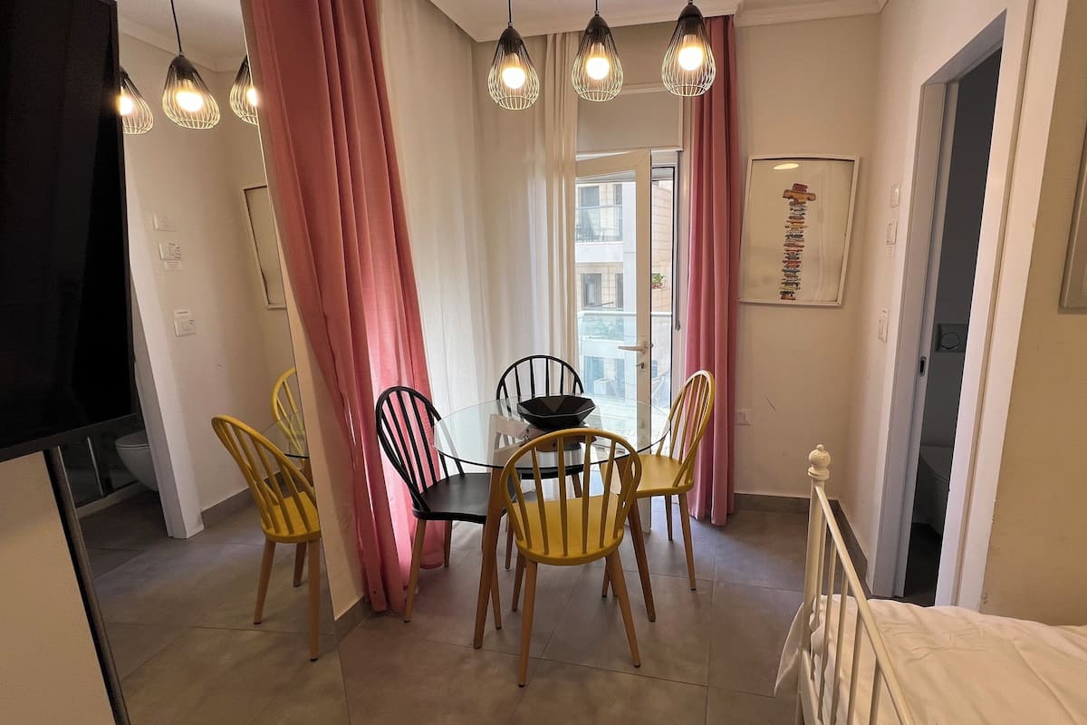 En matplats med ett glasbord, fyra stolar och en skål. Det finns rosa gardiner, en taklampa och en dörr med ett fönster.