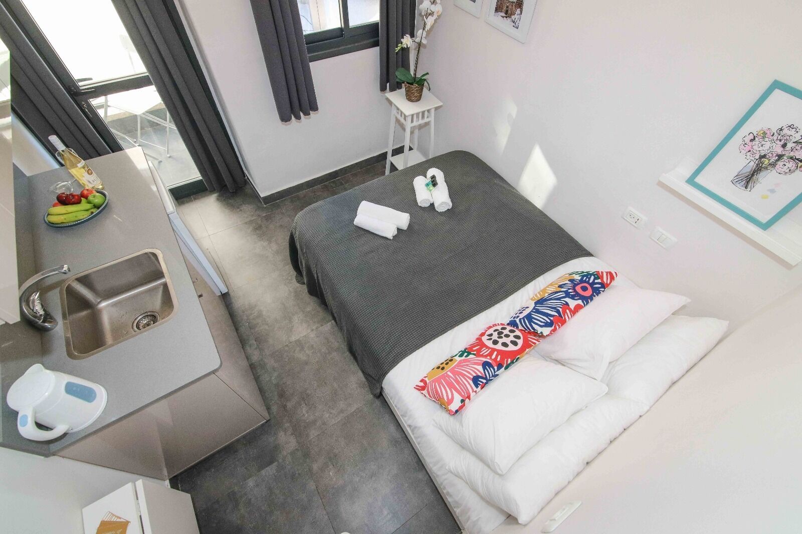 A hotel room with a bed, a small desk with a sink, a window with curtains, and a wall with a framed picture.