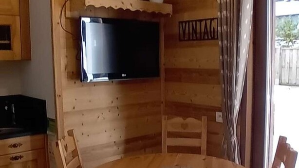 Interior de una cabaña de madera con una mesa de comedor, sillas, un televisor de pantalla plana montado en la pared y un ventanal con cortinas.