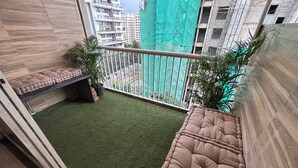 Un balcón con césped artificial, asientos acolchados, plantas en macetas y vistas de edificios circundantes.
