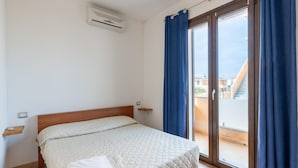 A bedroom with a bed, a window with curtains, a ceiling air conditioner, and a view of buildings outside. - Apartment in Residence With Private Terrace (Castelsardo)