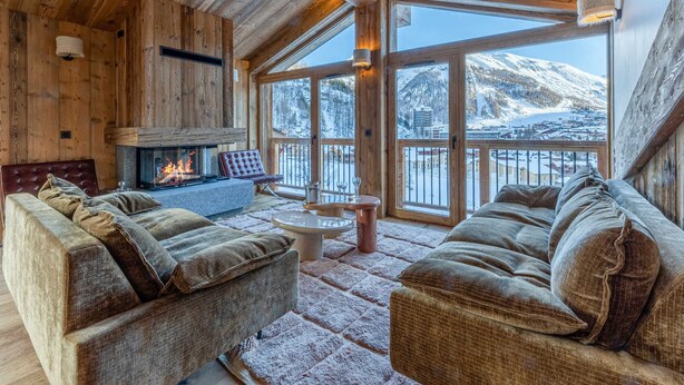 L’intérieur chaleureux d’un chalet en bois, avec une cheminée, des fauteuils confortables et une vue sur des montagnes enneigées.