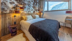 Une chambre avec un grand lit, une tête de lit en bois, un mur recouvert de fourrure, deux lampes de chevet, une fenêtre donnant sur la montagne et un fauteuil.