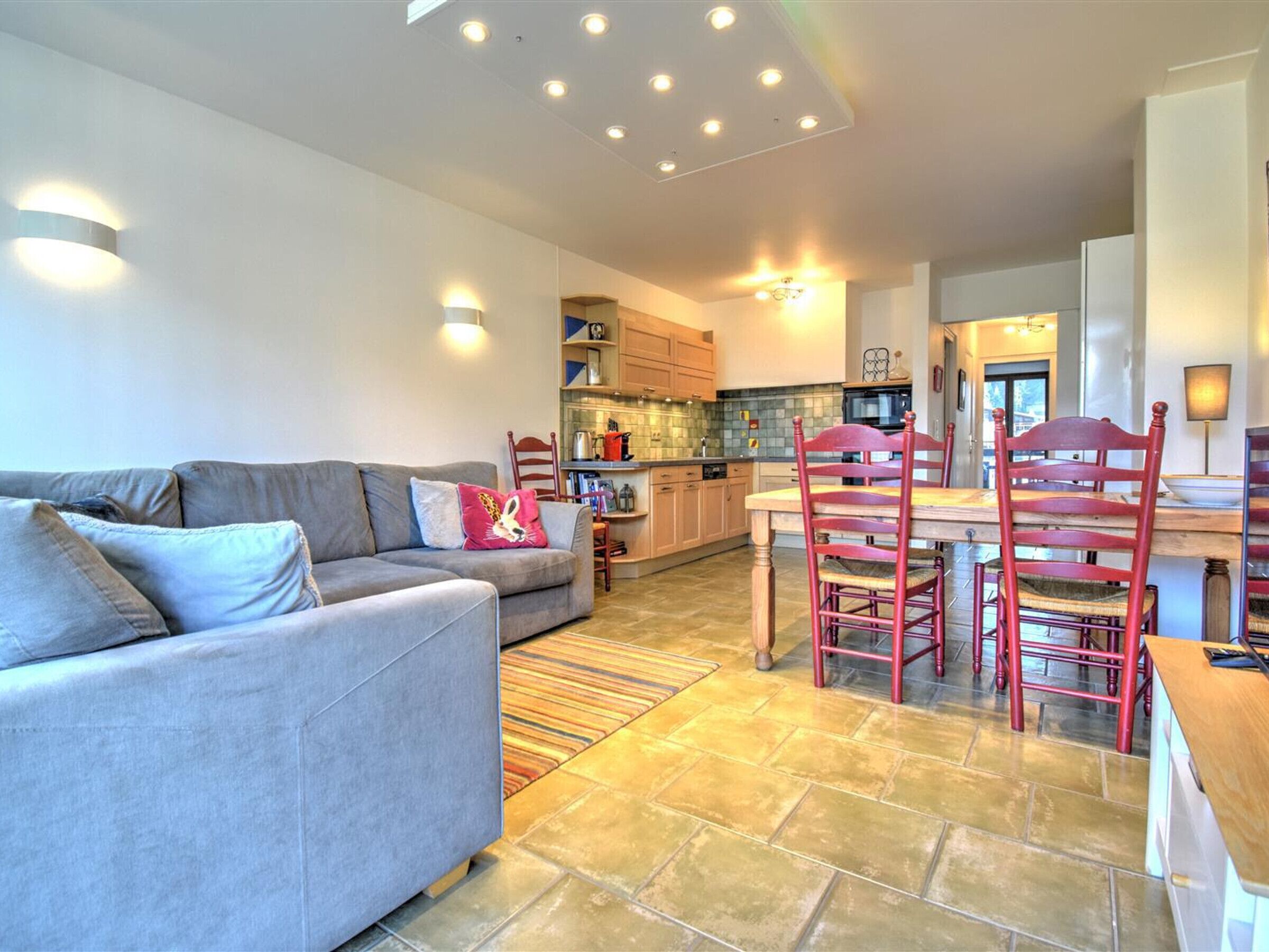 A modern living room with a grey sofa, a wooden dining table with red chairs, and a kitchen area with wooden cabinets and a microwave.
