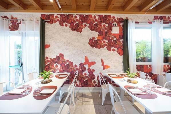 A dining area with white tables and chairs, a floral wall mural, and a view of greenery outside.