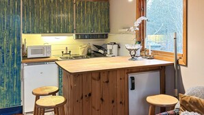 A compact kitchen with wooden cabinets, a microwave, a sink, and a small refrigerator. There are two stools and a window with a view of trees. - 4 Person Holiday Home in Tyreso (Tyreso)