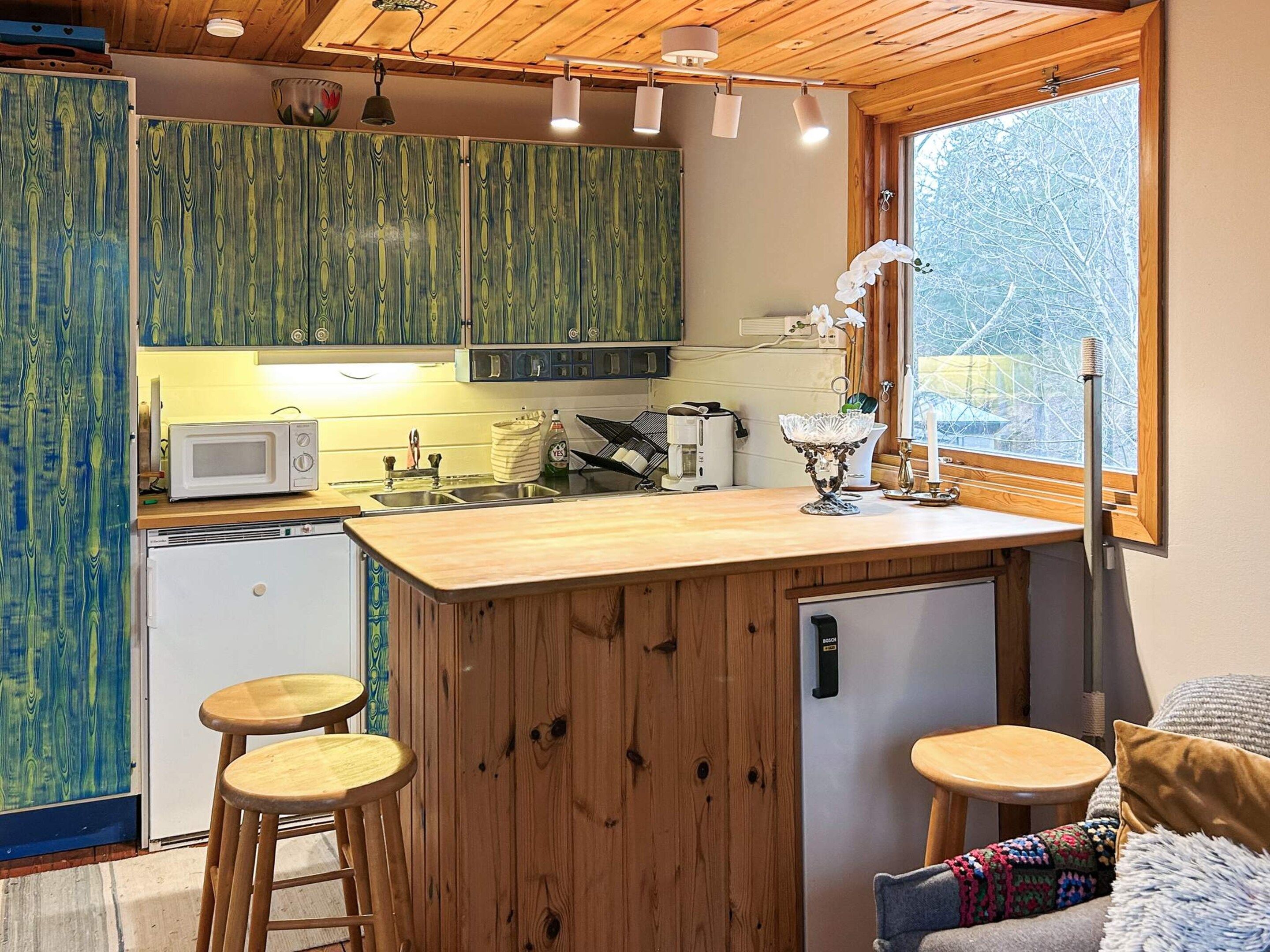 Cocina compacta con armarios de madera, un microondas, un lavamanos y un refrigerador pequeño. Hay dos taburetes y una ventana con vista a árboles.