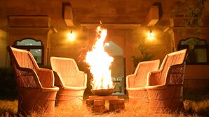 Wicker chairs arranged around a central fire pit with flames, set against a stone building facade. - Khushbu haveli hotel and restaurant (Pokhran)