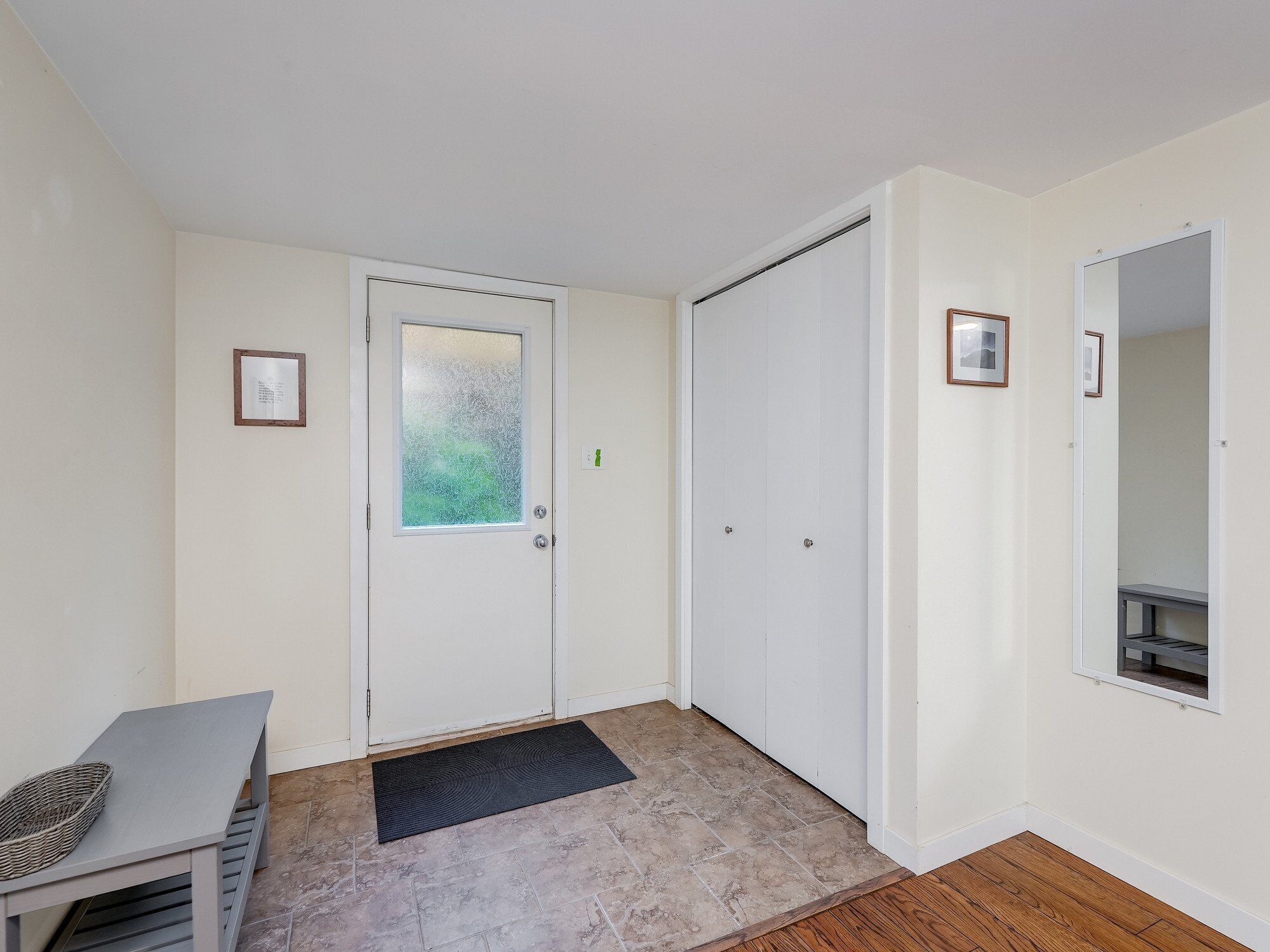 A room with a white door, a small table with a basket, a black doormat, and a wooden floor.