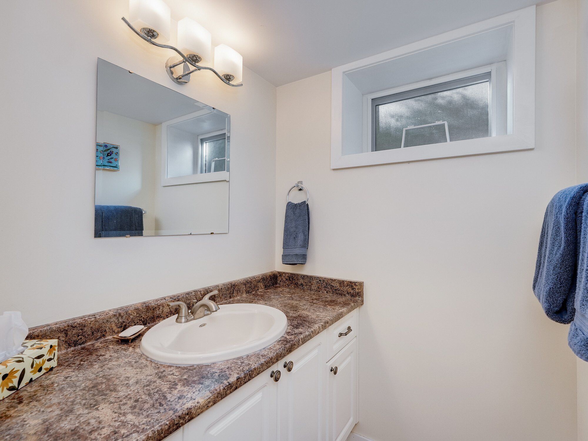 A bathroom with a granite countertop, white sink, and a large mirror. There is a window above the sink and a towel hanging on the wall.