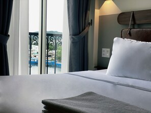 A hotel room with a bed, curtains, a view of boats and buildings through the window, and a wall-mounted light fixture. - PureJoy Hotel City Center Phu Quoc (Phu Quoc)