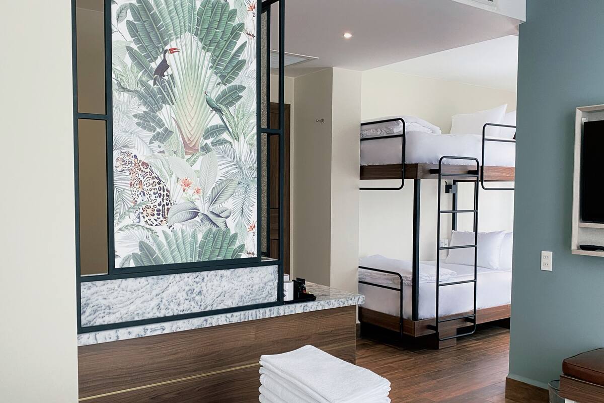 A hotel room with a bunk bed, a large window with a tropical print, a marble countertop, and a neatly stacked pile of towels.
