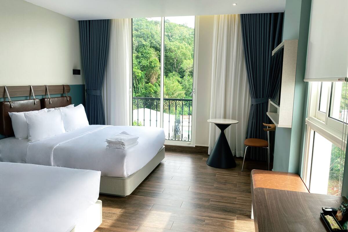 A hotel room with a large bed, a smaller bed, a desk, and a balcony with a view of greenery.