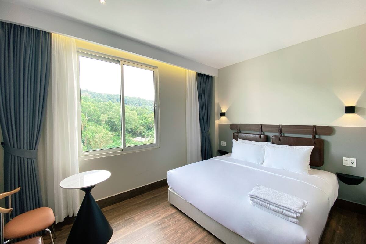 A hotel room with a large bed, a small round table, a chair, and a view of greenery through the window.