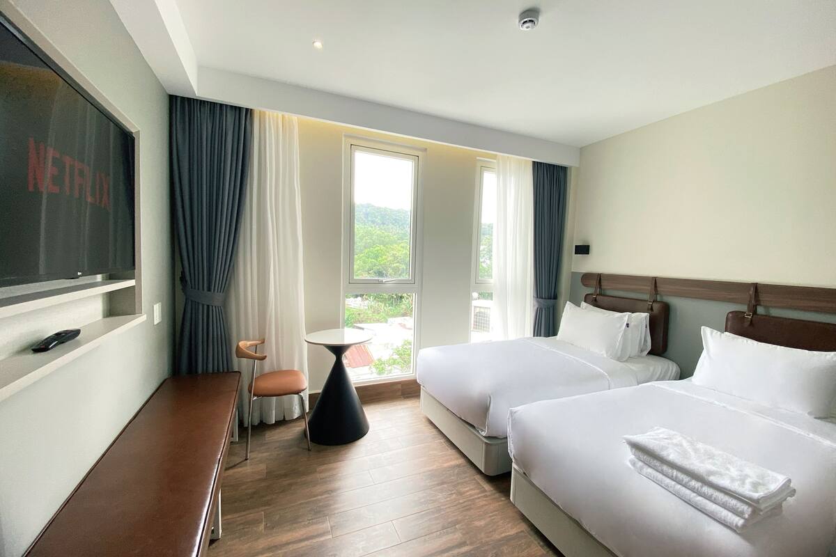 A hotel room with two beds, a TV, a small table, and a window with curtains.