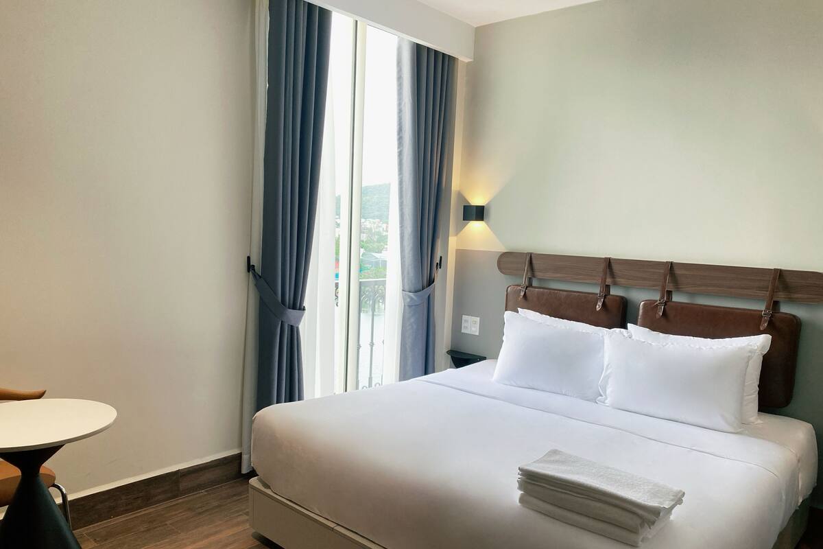 A hotel room with a bed, a nightstand, a table, and a window with curtains.