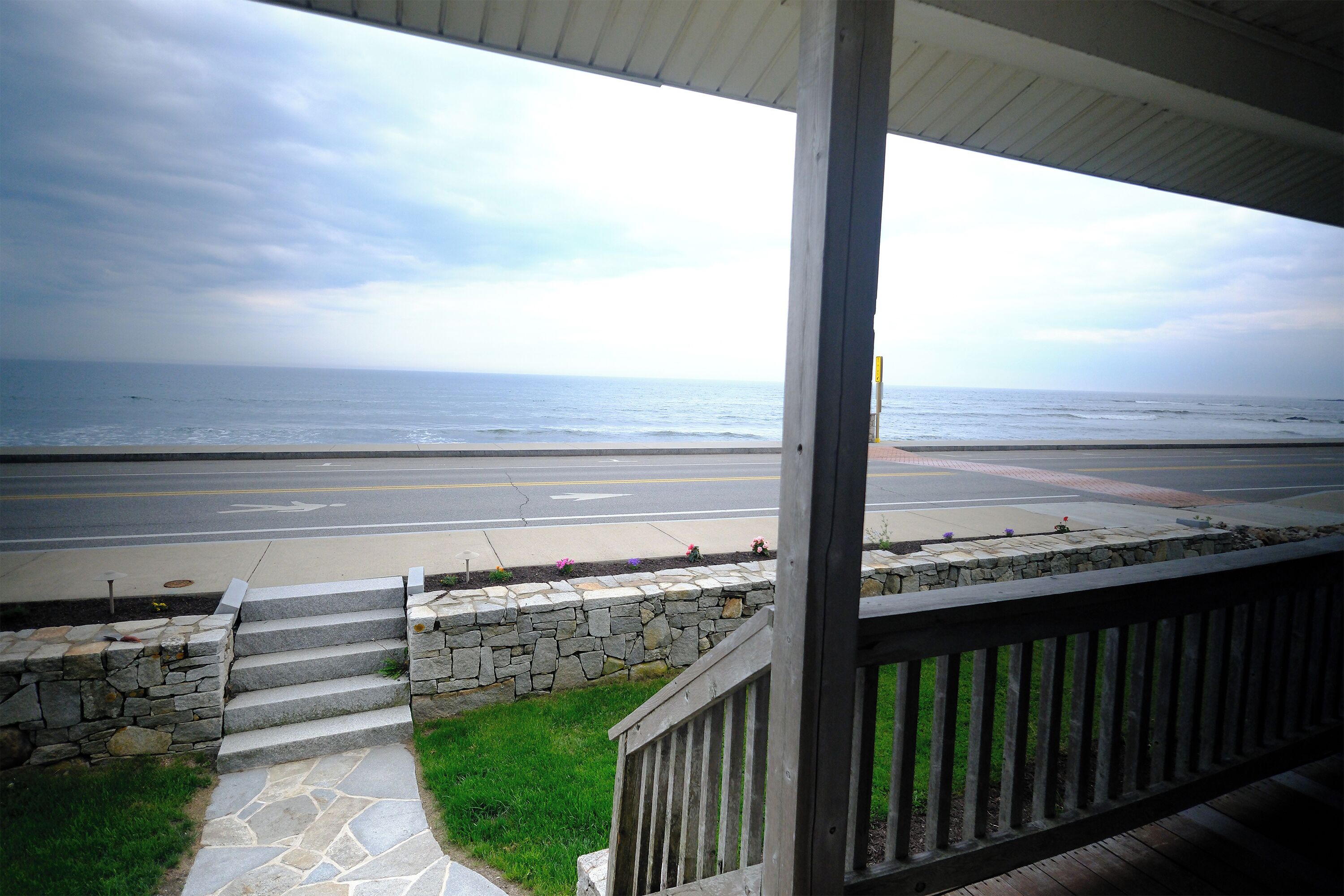 A view from a porch overlooking a road and beach, with stone steps leading down to the sidewalk.
