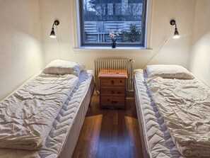 A bedroom with two single beds, a wooden dresser, a radiator, and a window with a view of the outside.