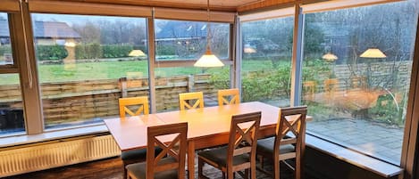 A dining area with a wooden table and chairs, large windows, and a view of a garden.