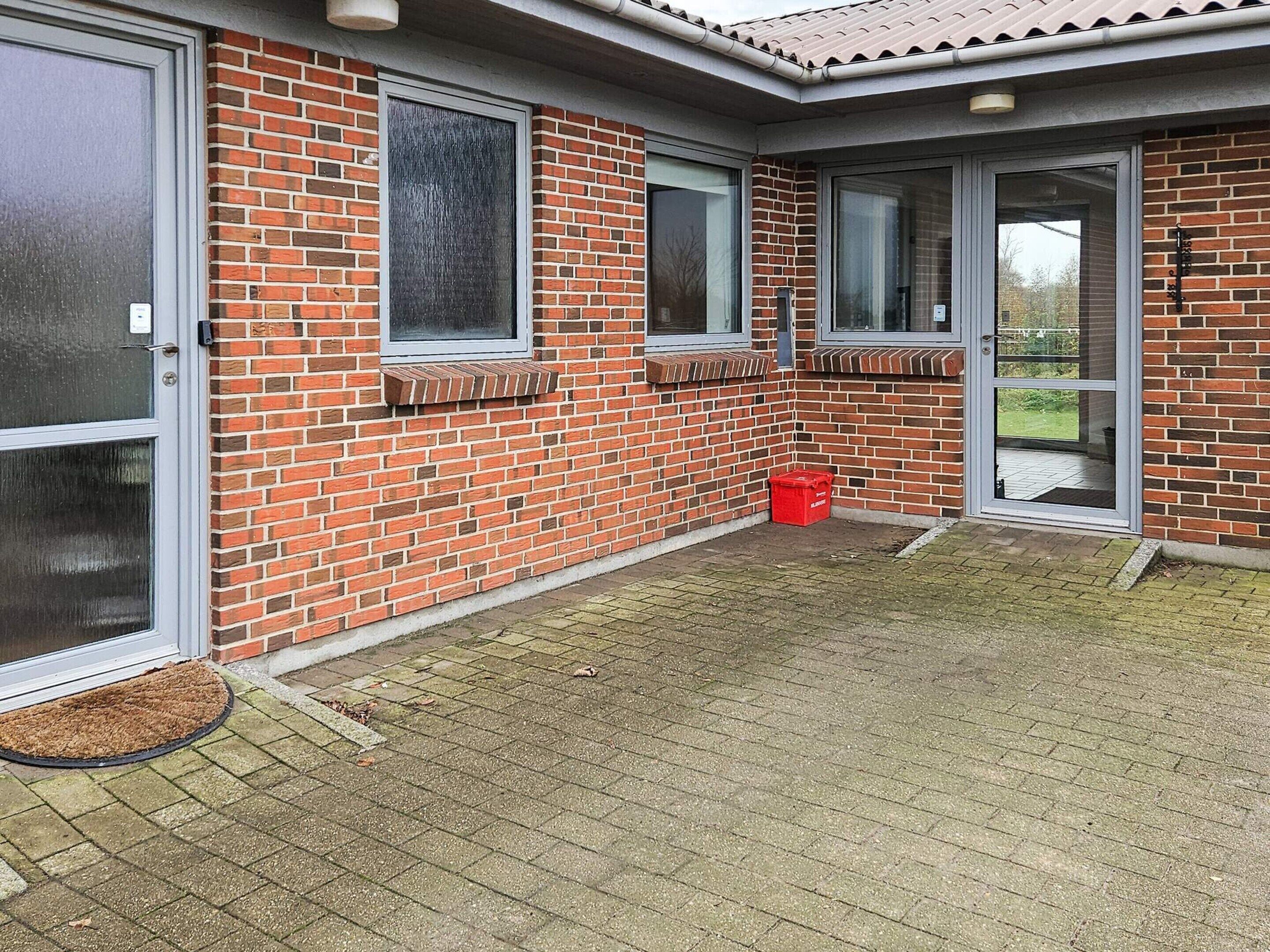 En murbygning med to store glasdøre og -vinduer, en rød affaldsbeholder og en dørmåtte.