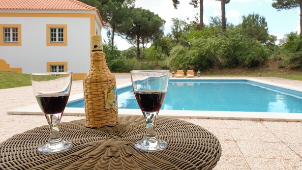 A wicker table with two wine glasses and a bottle, set against a poolside view with a building in the background. - UA 78- Villa dos Sobreiros (Alcácer do Sal)