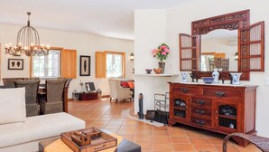A spacious living room with a sofa, a coffee table, and a chandelier. There is a dining area with a table and chairs, and a large wooden cabinet with a mirror. - UA 78- Villa dos Sobreiros (Alcácer do Sal)