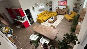Una sala moderna con un sofá amarillo, una mesa de centro de madera y un comedor con una mesa y sillas blancas. Hay plantas en macetas y un cuadro grande en la pared.