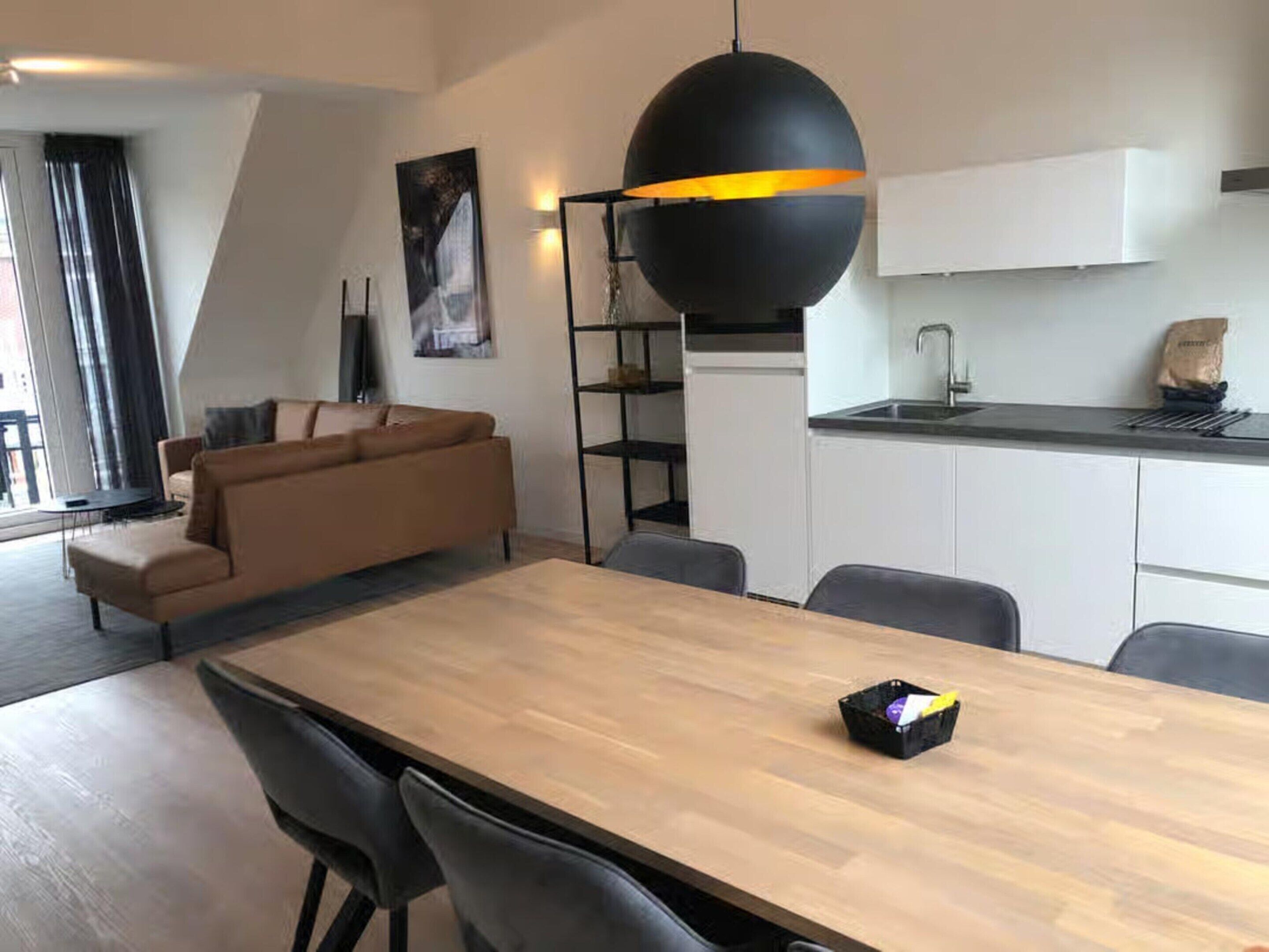 A modern living room with a wooden dining table, grey chairs, a brown sofa, a black pendant light, and a kitchen area with white cabinets and a black countertop.