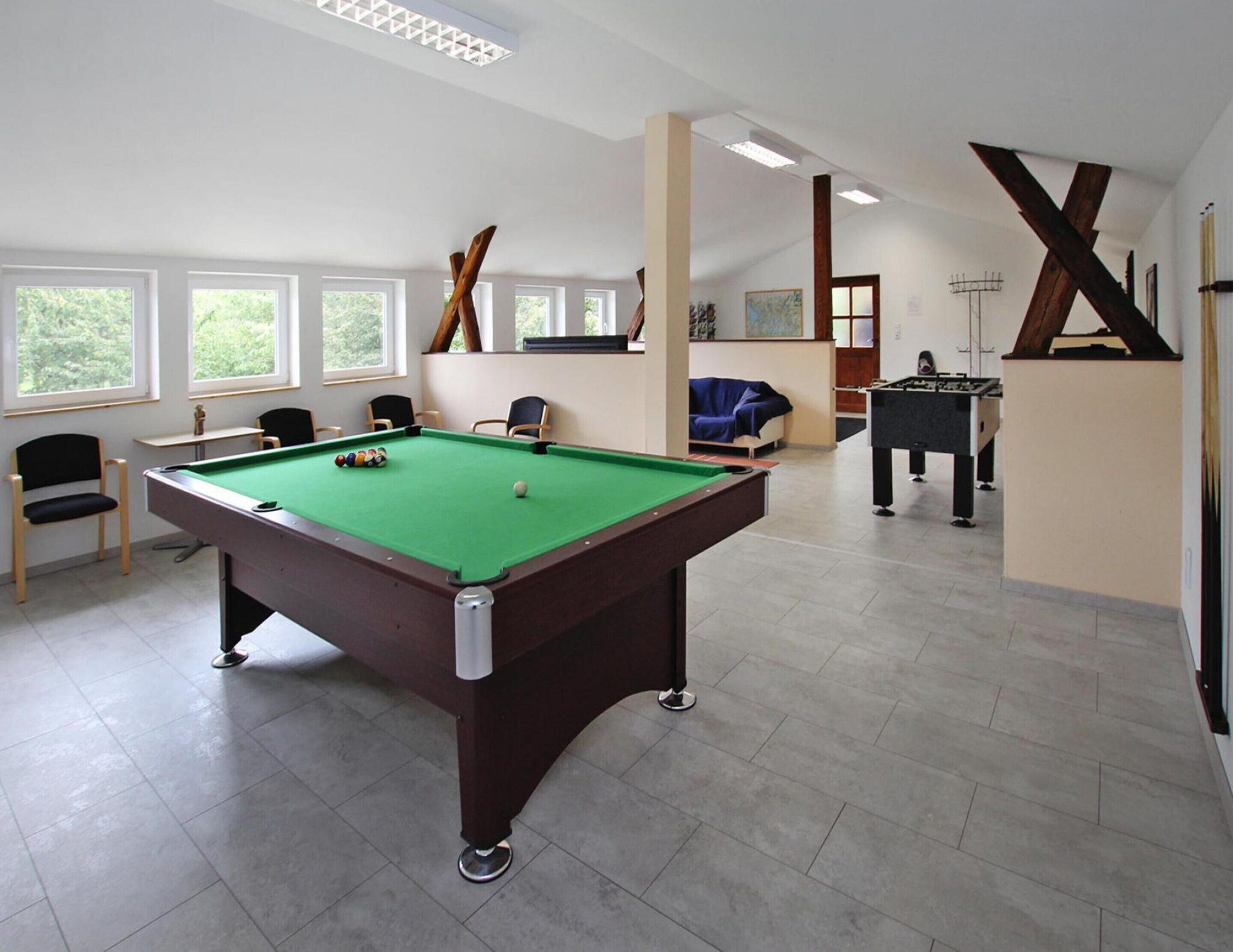 A pool table in a room with multiple windows, chairs, and a foosball table.