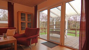Ein Zimmer mit Holzboden, einem roten Sofa, einem Glastisch und einer Glasschiebetür, die auf eine Terrasse im Freien führt.