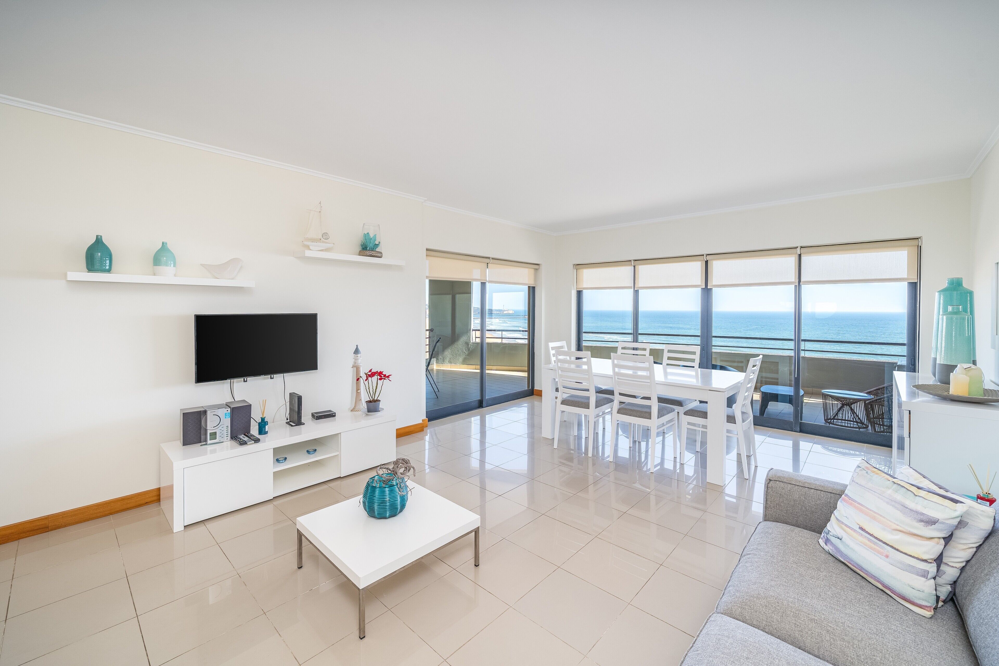 A modern living room with a flat-screen TV, white furniture, a dining area, and a view of the sea.