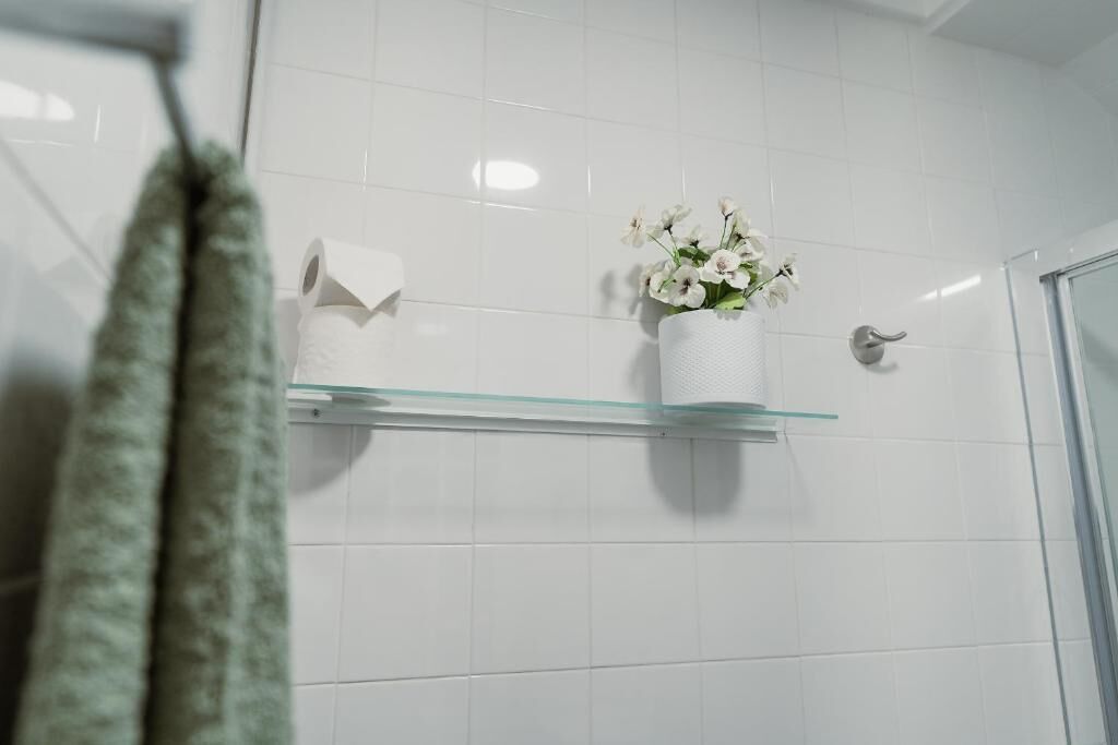Un bagno con piastrelle bianche, uno scaffale in vetro con un vaso di fiori e un asciugamano appeso al muro.