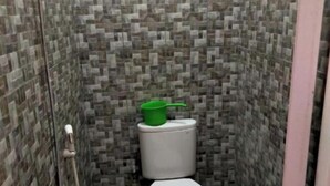 A small, tiled bathroom with a toilet, a green bucket on the tank, and a shower hose. - Homestay Komodo Mountain View (Batu Cermin)