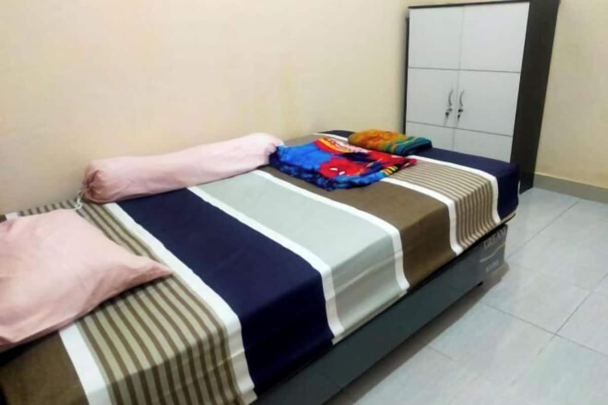 A neatly made bed with striped bedding and a colorful blanket, a pink pillow, and a wardrobe with two doors in a room with plain walls and tiled flooring.