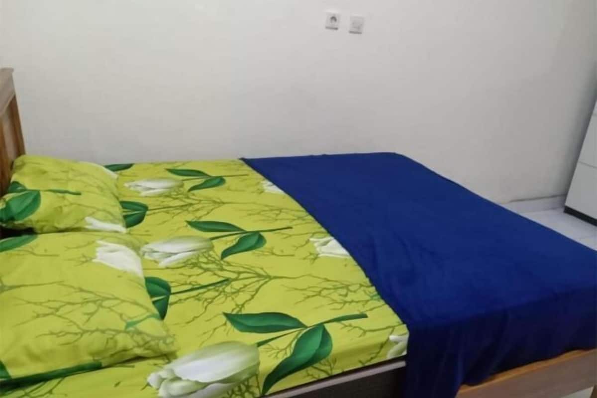 A bed with a yellow floral bedspread and blue blanket, a wooden headboard, and a white wall with two electrical outlets.