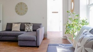 Un salon moderne avec un canapé d’angle gris, une horloge ronde fixée au mur et une plante en pot près de la fenêtre.