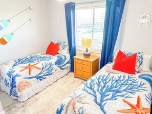 A bedroom with two beds, each with a starfish and coral themed bedspread, a nightstand with a lamp, and a window with curtains. - Tilghman Beach and Racquet Club 362 (North Myrtle Beach)