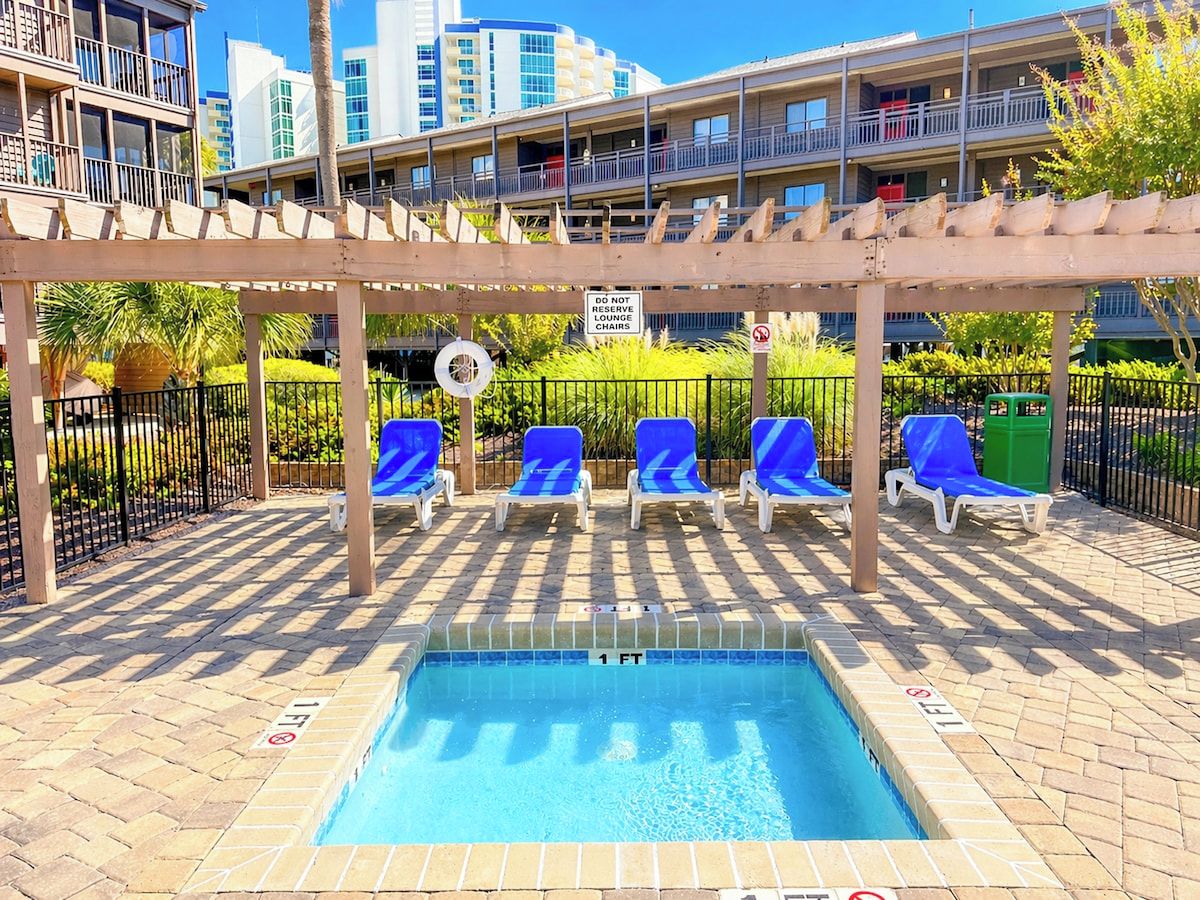 Ein Poolbereich mit Liegestühlen, einer Holzpergola und einem Schild, das eine Wassertiefe von etwa 30 Zentimetern angibt.