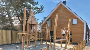 A wooden play structure in a sandy area with a house in the background. - Beautiful Lodge for 5-person Near the Beach (Zoutelande)