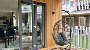 A wooden deck with a glass door leading to a kitchen, a hanging chair, and a white balcony railing. - Beautiful Lodge for 5-person Near the Beach (Zoutelande)