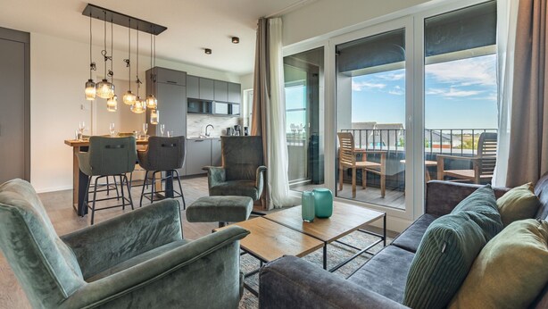A modern living room with a sofa, armchairs, and a coffee table. There is a dining area with chairs and a table, and a kitchen in the background. - Luxusapartement mit Meerblick (Nordstrand)