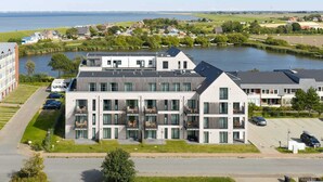 A modern multi-story building with balconies, a parking area, and a view of a lake and surrounding greenery. - Luxusapartement mit Meerblick (Nordstrand)