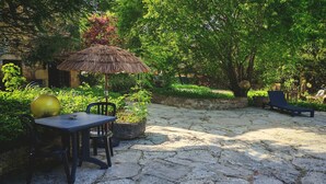 Une terrasse en pierre avec une table et des chaises, un parasol en chaume et un fauteuil de relaxation.