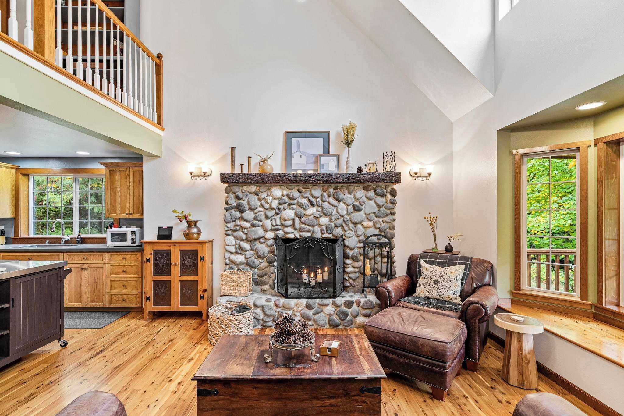 A spacious living room with a stone fireplace, wooden flooring, a leather sofa, and a high ceiling with a balcony.