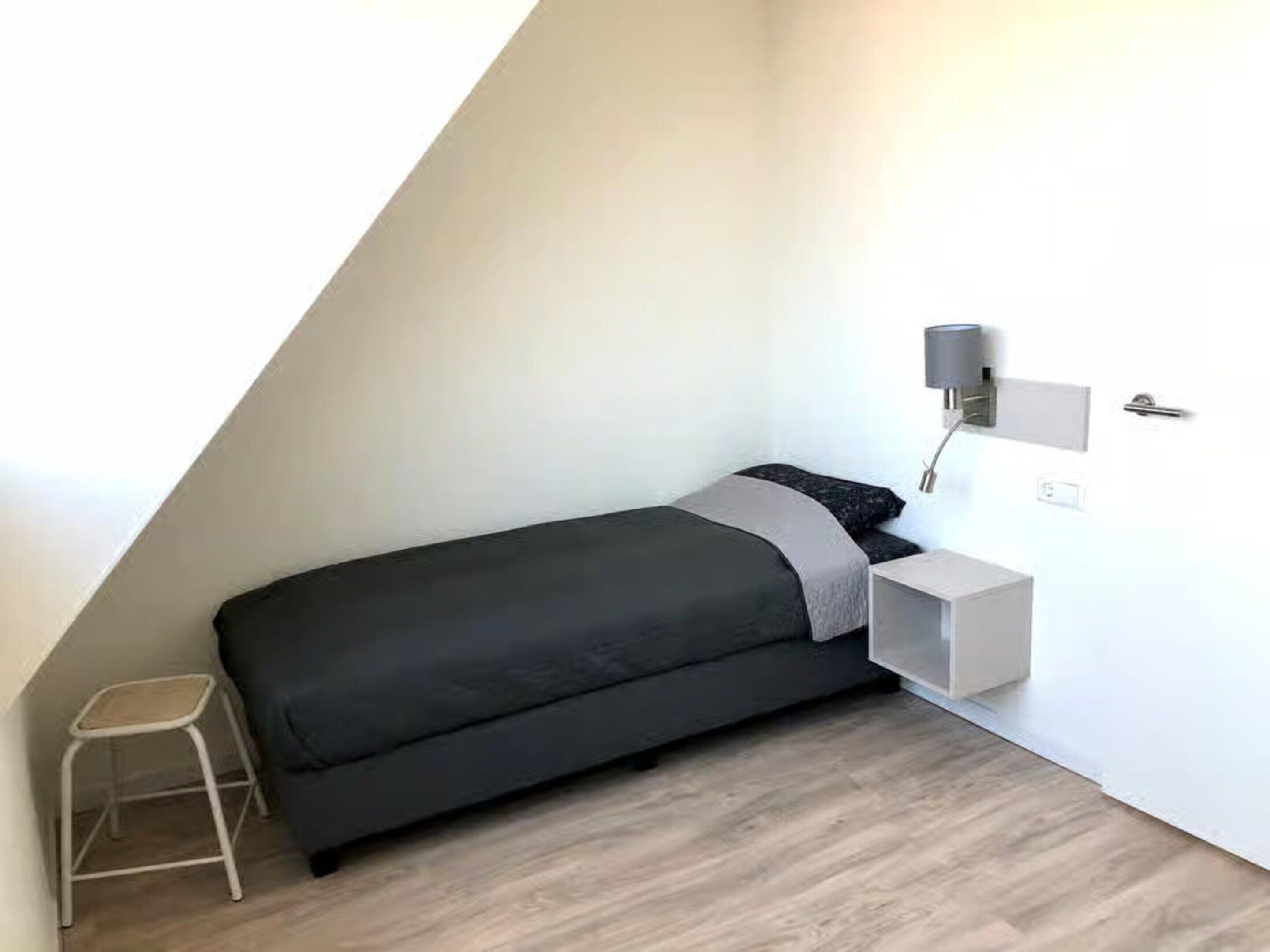 Une petite chambre à coucher moderne, avec un lit, une table de chevet, une lampe et un tabouret.
