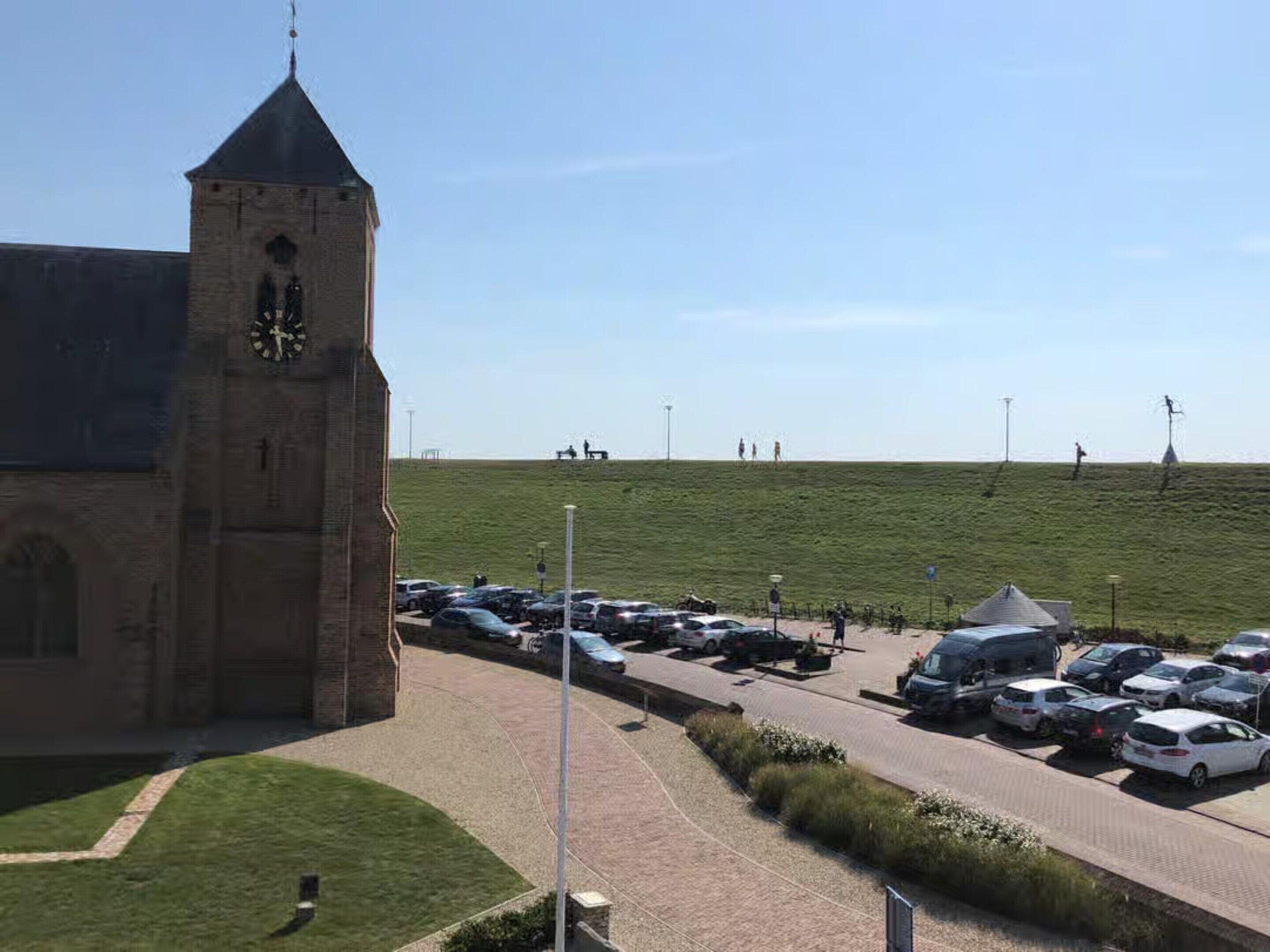 Une église dotée d’un clocher, un parking et un champ verdoyant où l’on aperçoit des personnes au loin.