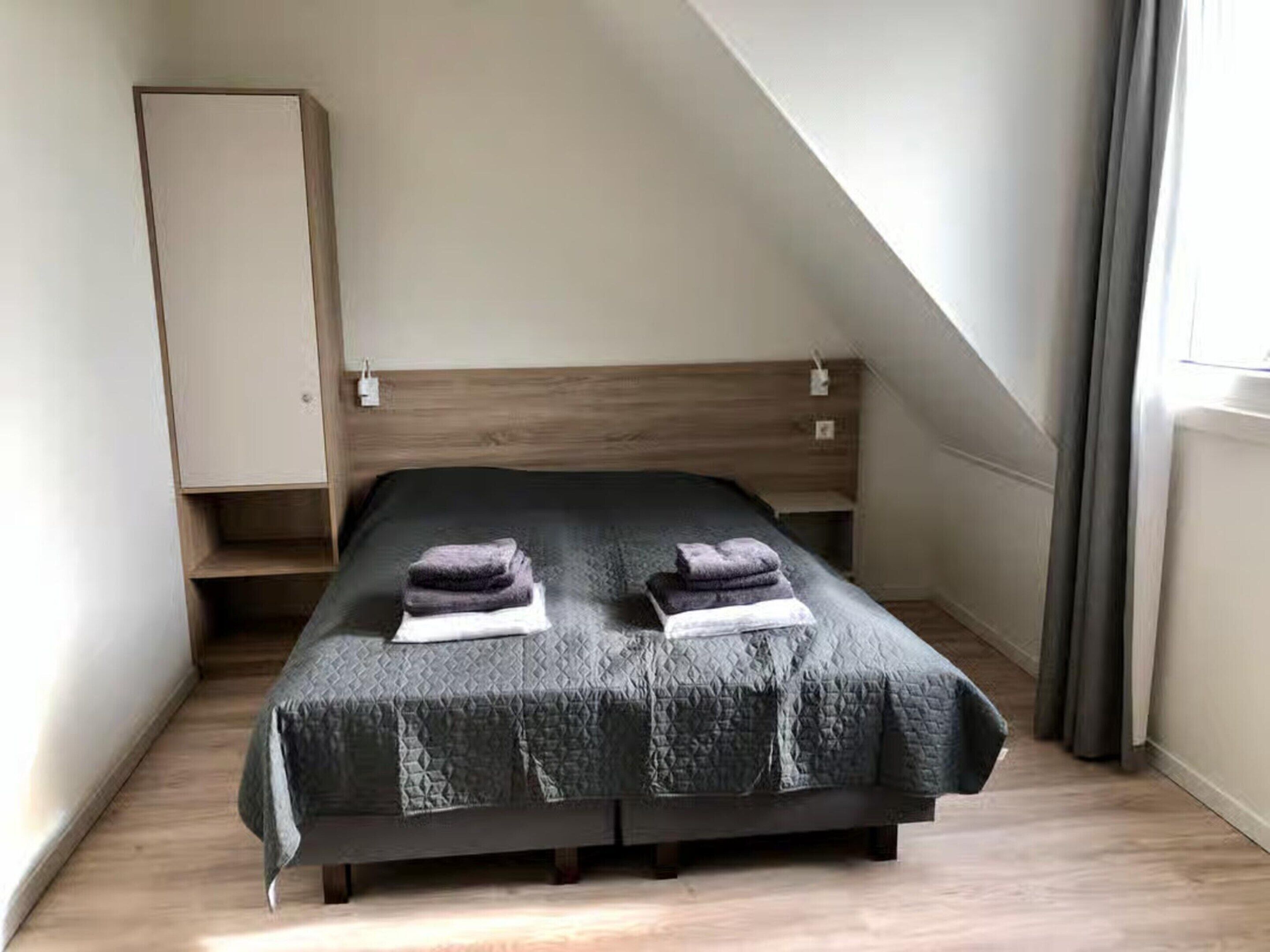 A single bed with a dark quilted bedspread and two folded towels on top.