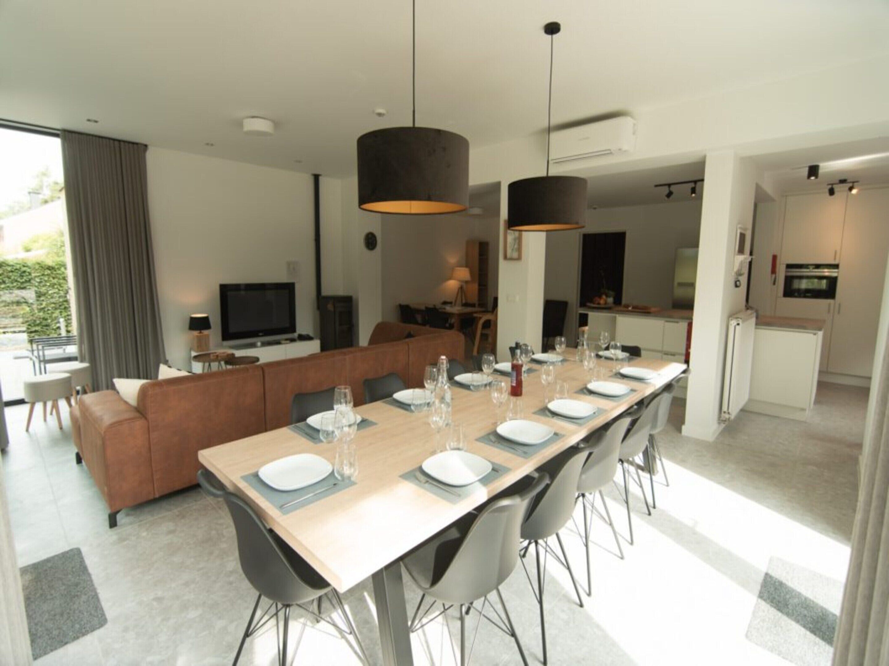 Un espace de repas moderne avec une longue table dressée pour six personnes, un canapé et un téléviseur en arrière-plan.