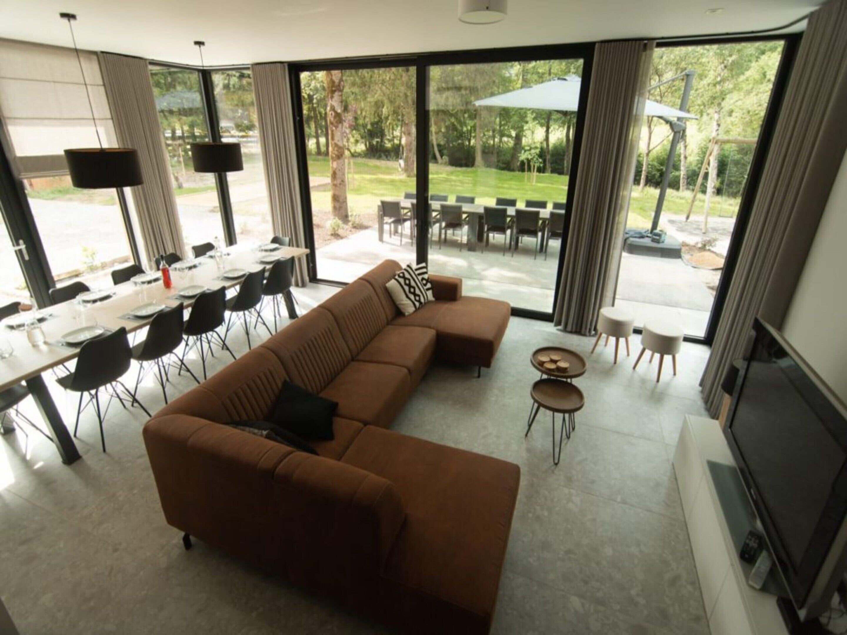A modern living room with a brown sectional sofa, a dining area with a table and chairs, and a large sliding glass door leading to an outdoor patio.